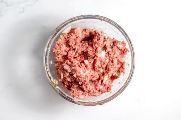 A glass bowl filled with a mixture of ground beef, chopped onions, and seasoning sits on a white surface.