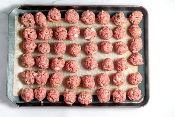 A baking sheet lined with parchment holds rows of raw, evenly spaced meatballs mixed with herbs and chopped onions.