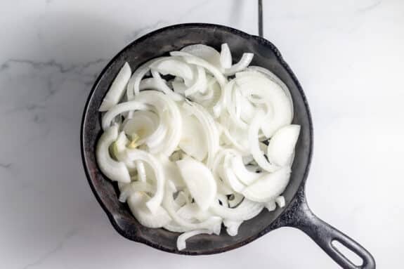 A cast iron skillet filled with raw, sliced white onions on a white marble surface.