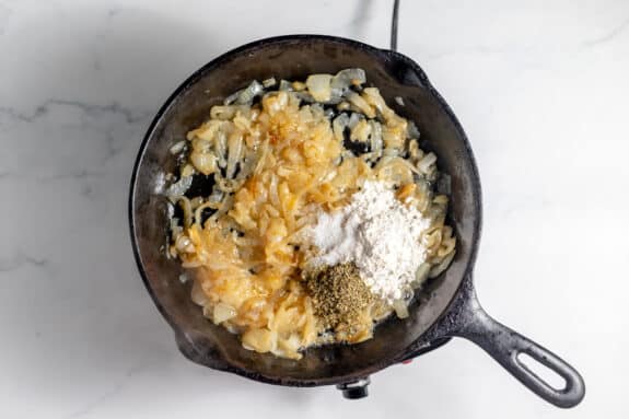 A cast iron skillet on a stovetop contains sautéed onions, flour, and herbs on a white marble surface.