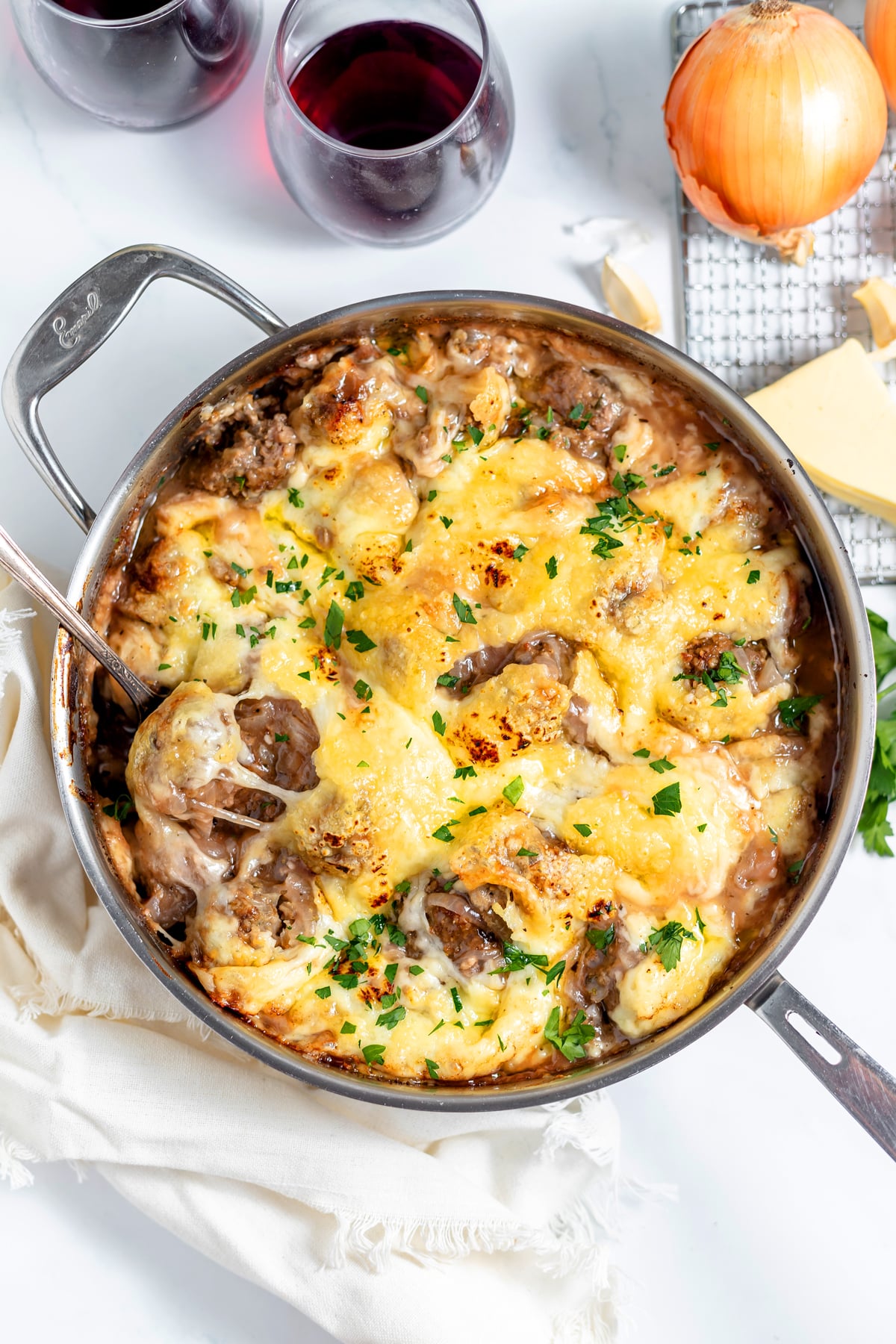 A skillet filled with french onion meatballs with ground beef and potato casserole, garnished with parsley, next to an onion, cheese, and two glasses of red wine.