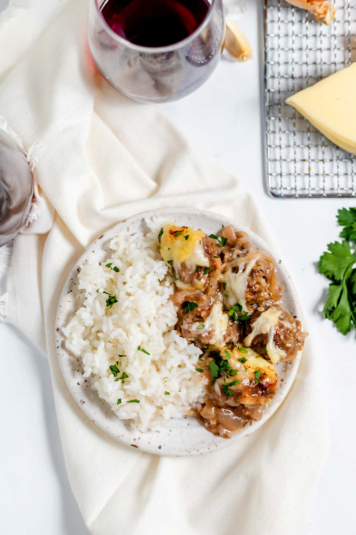 A plate with white rice and french onion meatballs in sauce, garnished with herbs, next to a glass of red wine, cheese, and fresh parsley on a light-colored table.