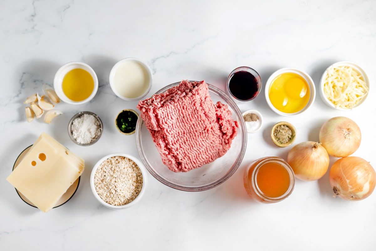 A variety of french onion meatballs ingredients, including Lean ground beef (85/15), milk, quick oats, parsley, Italian seasoning, salt, black pepper, garlic, eggs, olive oil, yellow onions, dry red wine, chicken broth, flour, Swiss cheese, Gruyere cheese, arranged on a white surface in bowls and small dishes.