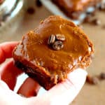 A hand holding a chocolate brownie topped with smooth chocolate frosting and two whole coffee beans.