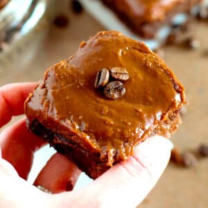 A hand holding a chocolate brownie topped with smooth chocolate frosting and two whole coffee beans.