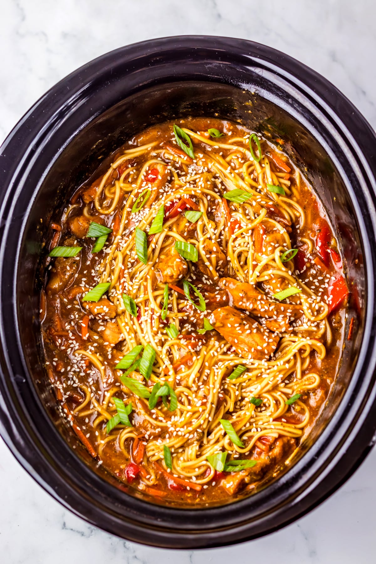 A Slow Cooker Pork Lo Mein recipe filled with cooked noodles, vegetables, and sauce, topped with chopped green onions and sesame seeds.