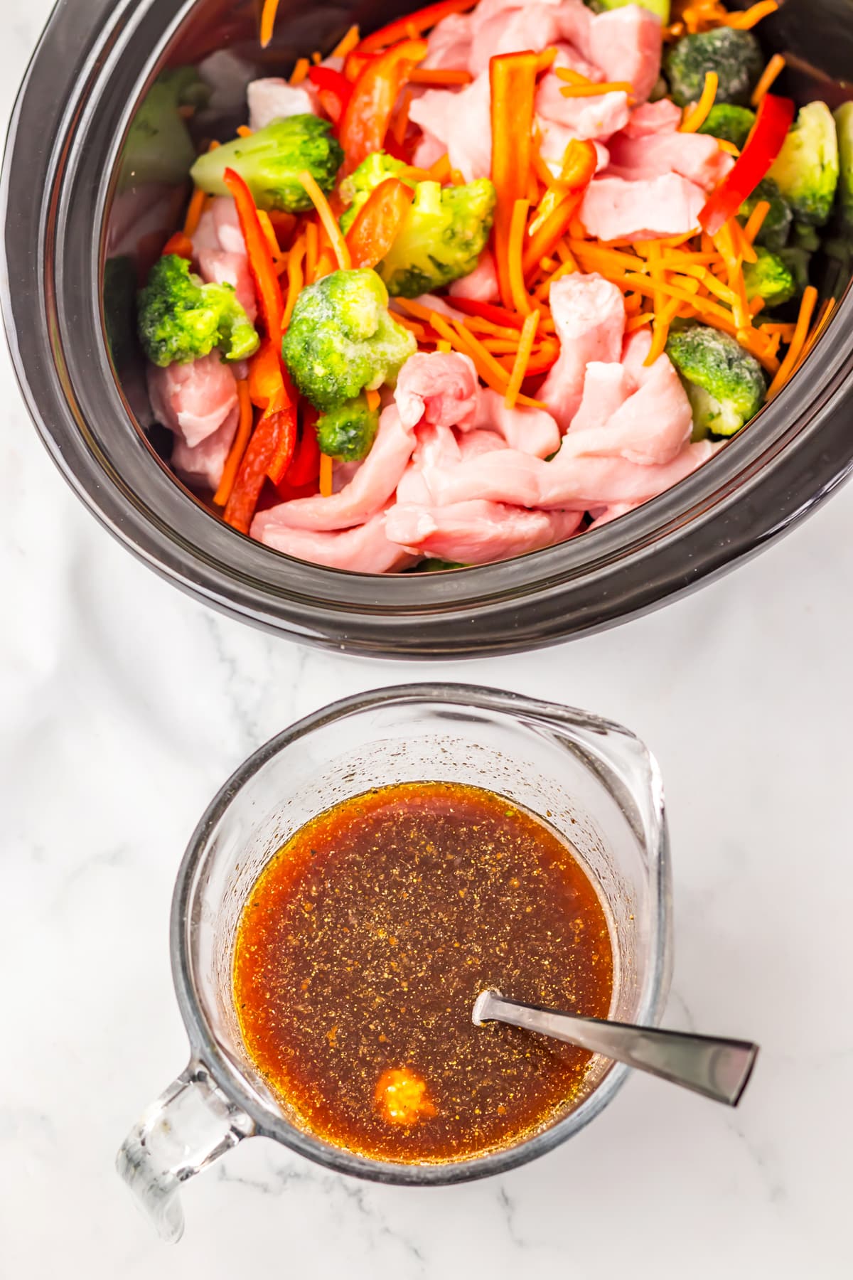 First step in preparing Slow Cooker Pork Lo Mein includes preparing the sauce by whisking together soy sauce, chicken stock, rice vinegar, hoisin sauce, sesame oil, garlic powder, ground ginger, black pepper, ketchup, and minced garlic. Sliced pork, broccoli, carrots, and red bell pepper in a slow cooker; a glass measuring cup with brown sauce and a spoon sits beside it on a white countertop.