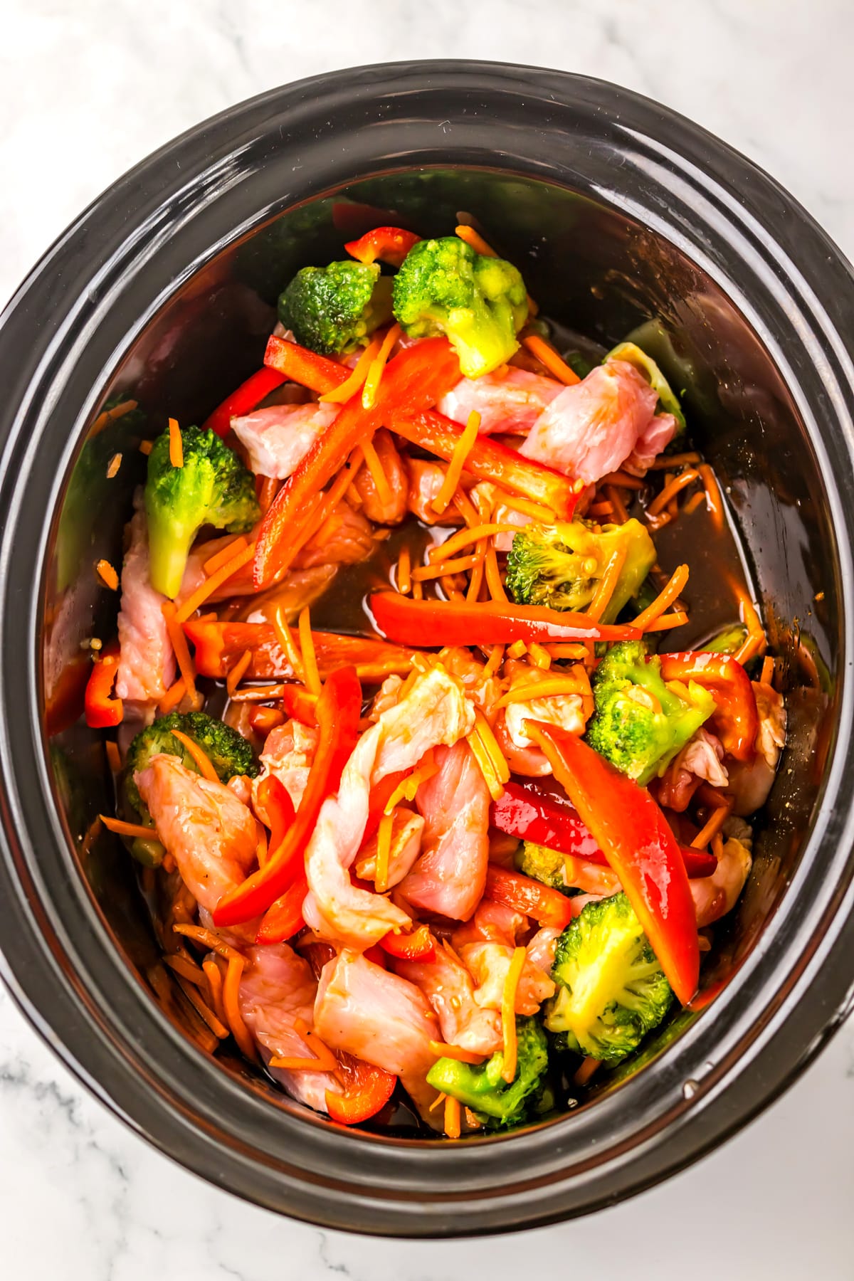 Next step in preparing Slow Cooker Pork Lo Mein is to add the sauce to the slow cooker with uncooked pork strips, broccoli, red bell peppers, and carrots on a marble surface.