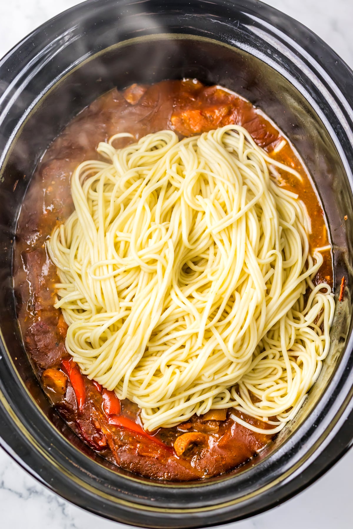 Adding lo mein noodles on top of mixture with vegetables in a black slow cooker, with steam rising from the hot food.