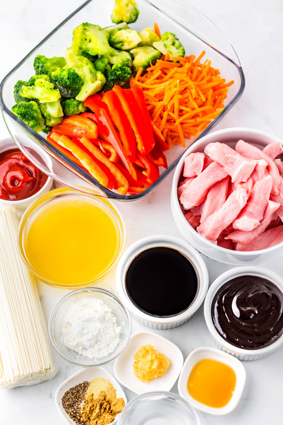 Assorted stir fry ingredients for Slow Cooker Pork Lo Mein includes: broccoli, carrots, bell peppers, raw chicken, noodles, garlic powder, hoisin sauce, ground ginger, black pepper, ketchup, pork chops, shredded carrots,red bell peppers, cornstarch, water, Lo mein noodles, sesame seeds, and chicken stock arranged on a white surface.