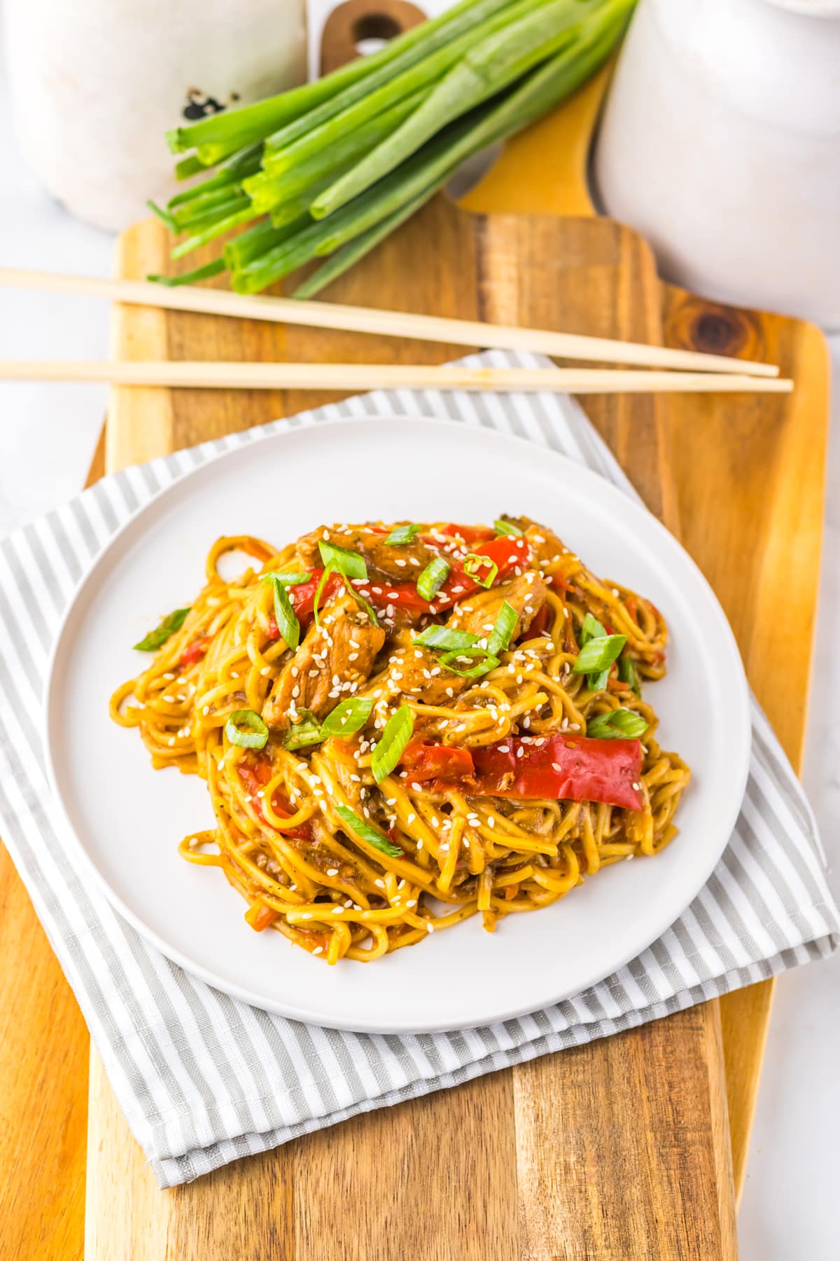 A plate of Slow Cooker Pork Lo Mein with sliced red bell peppers and green onions, garnished with sesame seeds, placed on a striped napkin with chopsticks and scallions nearby.
