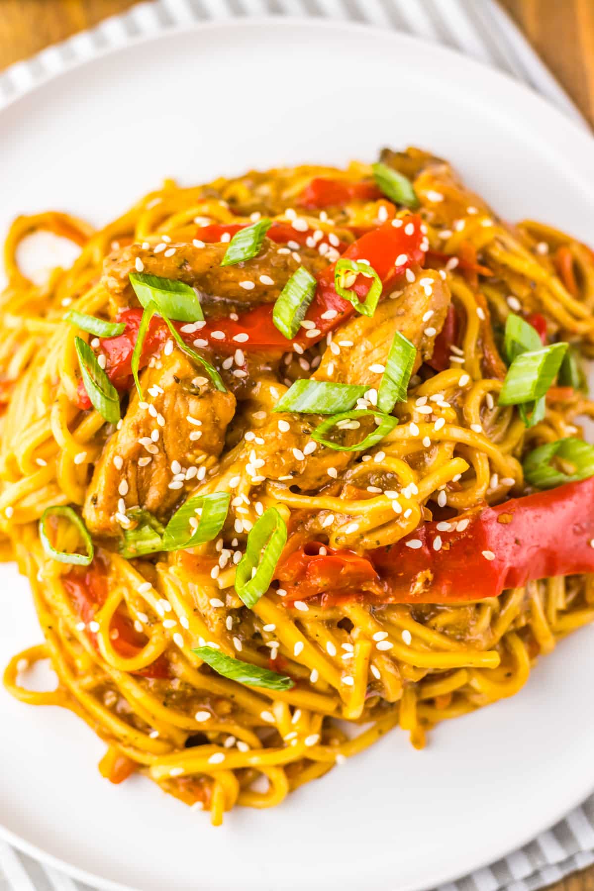 A plate of stir-fried noodles with sliced chicken, red bell peppers, green onions, sesame seeds, and sauce, served on a white dish.