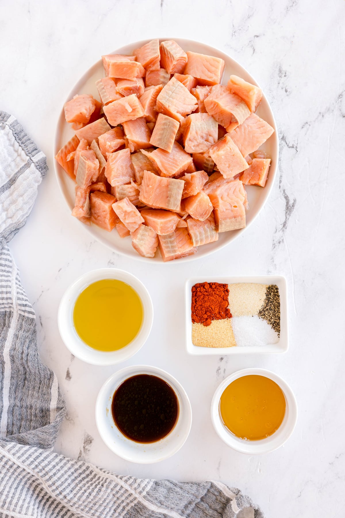 Ingredients for Air Fryer Salmon Bites are the following: a plate of cubed salmon with small bowls of olive oil, soy sauce, honey, and smoked paprika, garlic powder, onion powder, salt, black pepper, on a marble surface next to a striped kitchen towel.