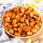 A plate of cooked, seasoned air fyer salmon bites is displayed on a wooden board, surrounded by lemon wedges and a striped cloth napkin.
