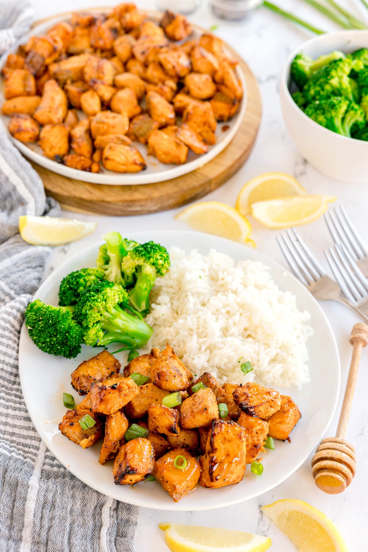 A plate with air fryer salmon bites with white rice, and steamed broccoli, with extra chicken, lemon wedges, and a bowl of broccoli in the background.