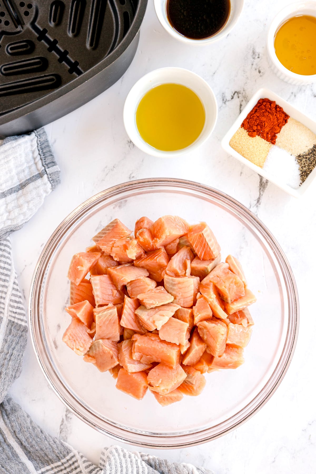 A glass bowl of raw salmon cubes dried with paper towels sits on a marble counter near an air fryer, with small bowls of oil, sauce, and seasonings arranged nearby. This is for Air Fryer Salmon Bites recipe.