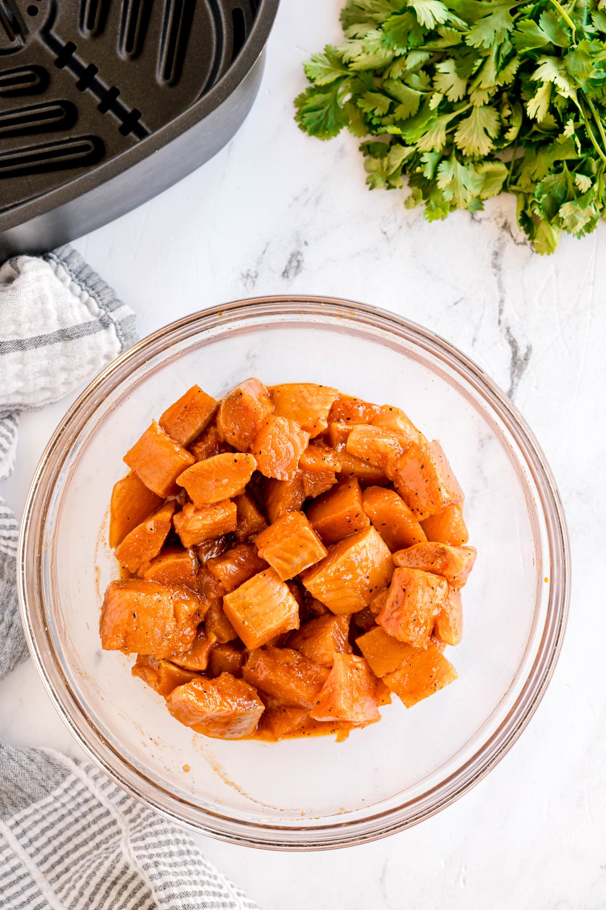 Next step in preparing air fyer salmon bites is pouring the marinade over the salmon and toss gently to coat. Seasoned, cubed raw chicken sits on a white countertop near fresh cilantro, a striped towel, and part of an air fryer basket.