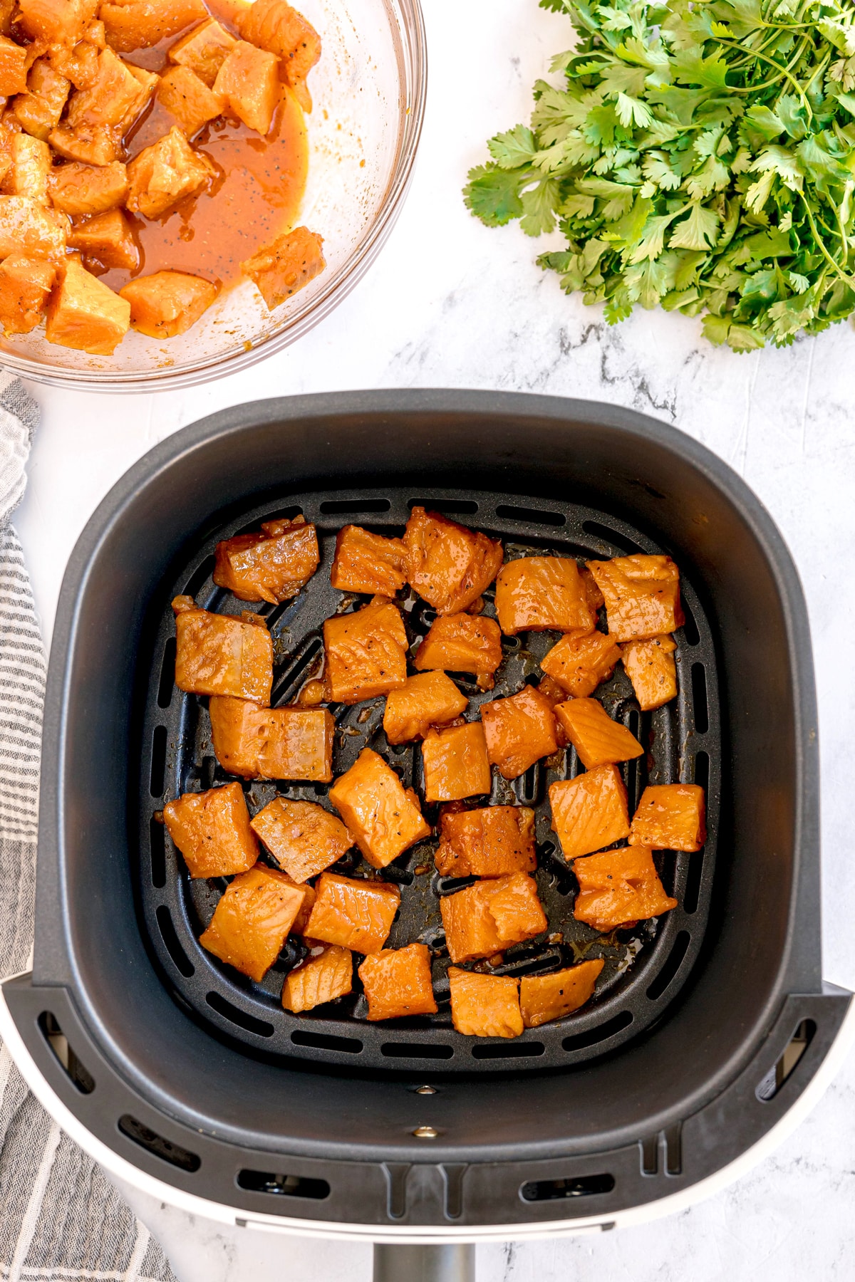 Next process in making air fryer salmon bites is to add seasoned sweet salmon in air frye basked. A bowl of seasoned salmon cubes and fresh cilantro visible nearby.