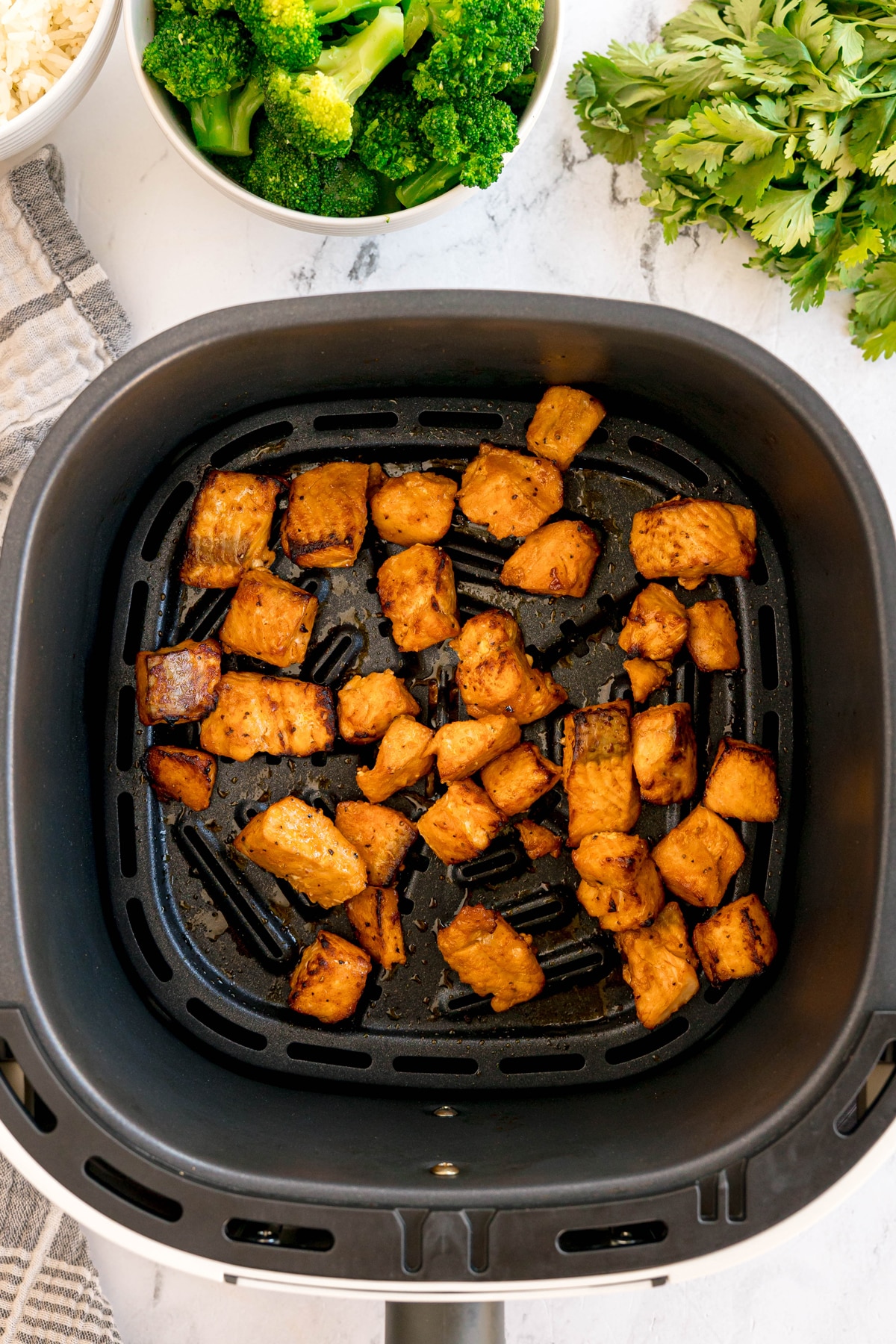 Freshly-cooked, seasoned Air Fryer Salmon Bites inside an air fryer basket, with bowls of broccoli and fresh herbs on the side.
