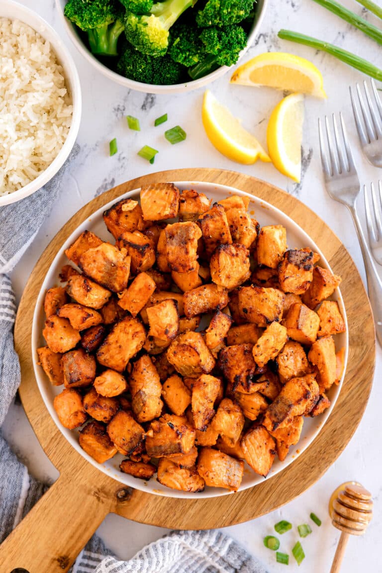A plate of seasoned Air Fryer Salmon Bites, on a wooden board, with bowls of rice and broccoli, lemon wedges, green onions, and forks nearby.