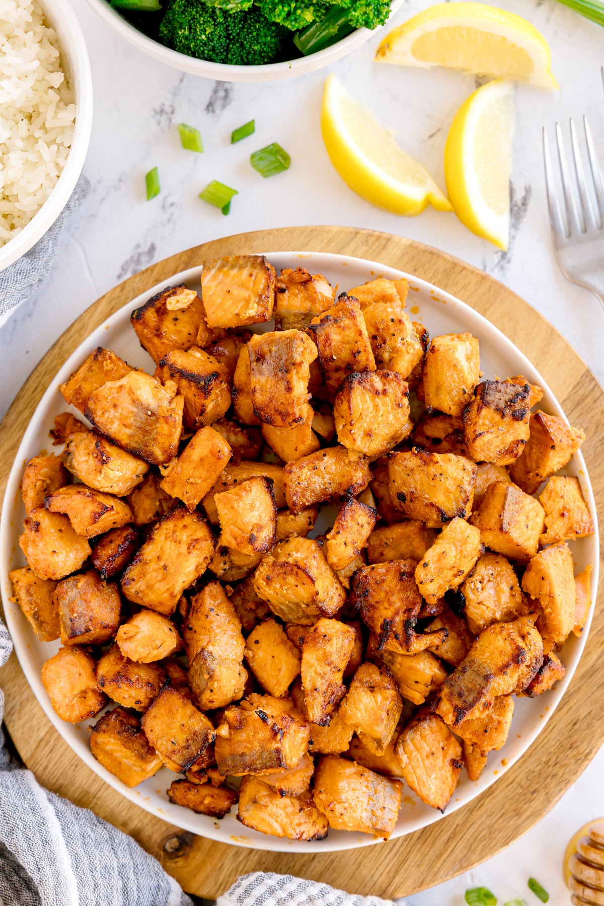 A plate of seasoned, air fryer salmon bites sits on a wooden board, surrounded by rice, broccoli, lemon wedges, and green onion garnish.