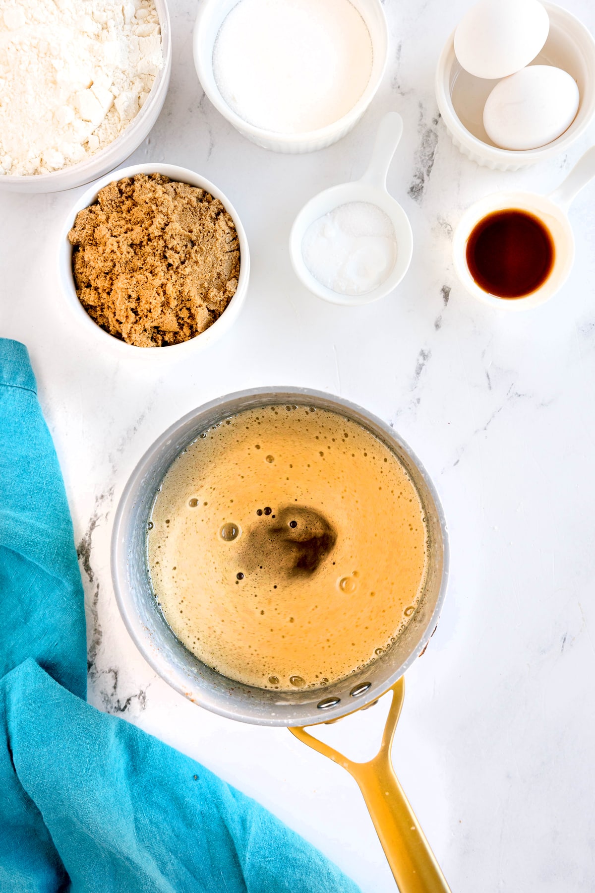 A saucepan with melted butter sits on a marble surface, surrounded by bowls of flour, sugar, brown sugar, baking soda, eggs, and vanilla extract. A blue cloth is beside them. This is the first process in preparing Brown Butter Cookies