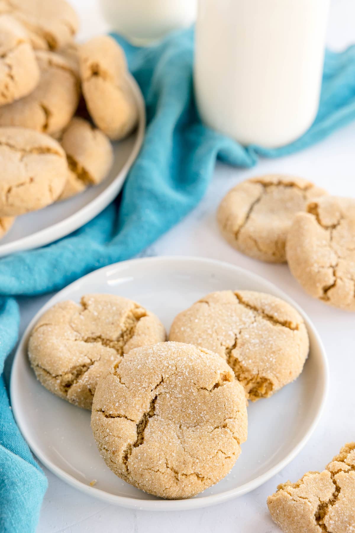 Brown Butter Cookies on a white plate, with more cookies and a bottle of milk in the background, set on a white surface with a blue cloth.