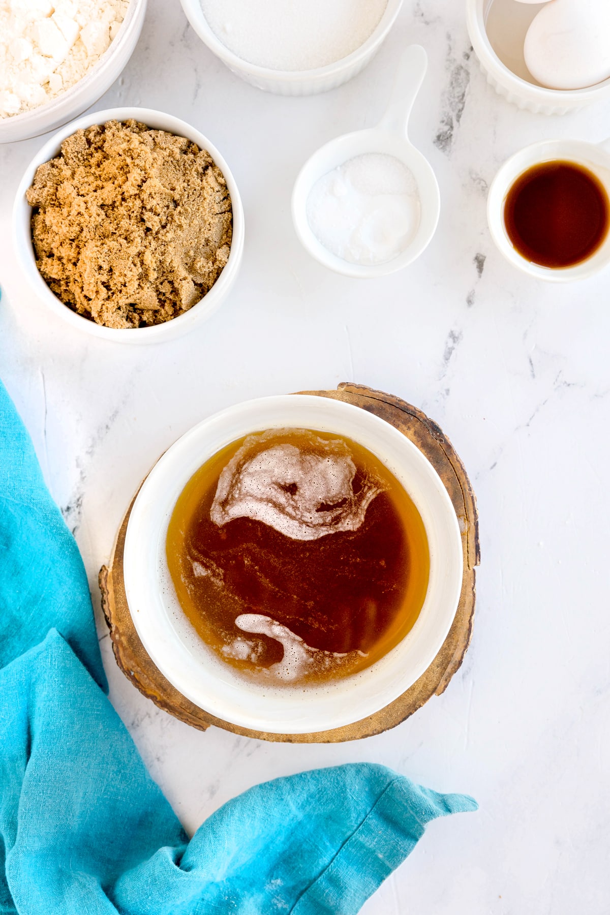 A bowl of melted butter on a wooden coaster surrounded by bowls of brown sugar, white sugar, flour, baking powder, and vanilla on a white surface with a blue cloth. This is one of the steps in preparing Brown Butter Cookies