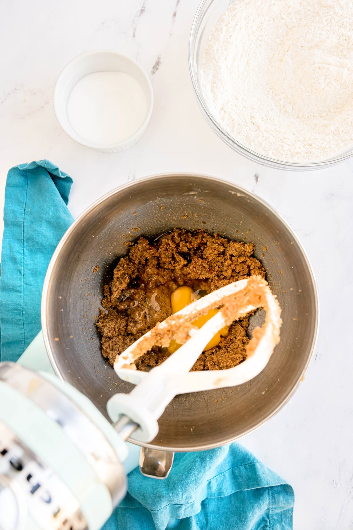 A mixing bowl with brown batter and an egg, a mixer paddle, a bowl of flour, and a small bowl of sugar on a white countertop with a blue cloth. This is one process in preparing Brown Butter Cookies