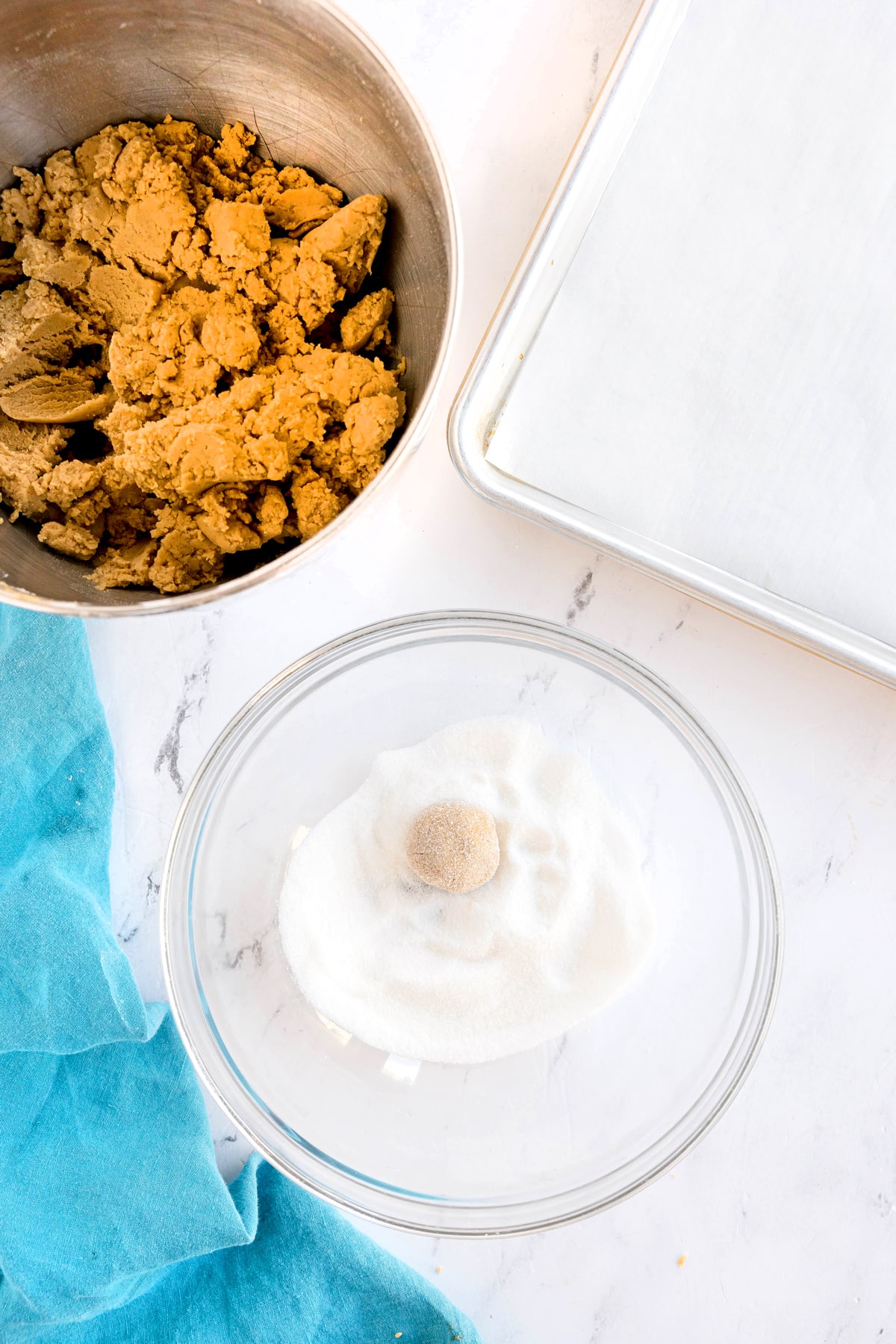 A bowl of dough, for Brown Butter Cookies a glass bowl with a dough ball in sugar, and an empty parchment-lined baking sheet on a marble surface with a blue cloth.