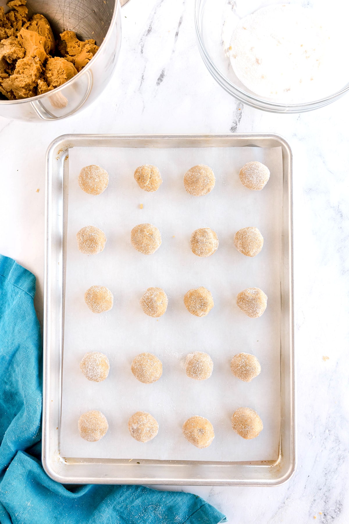 A baking tray lined with parchment paper holds evenly spaced, unbaked Brown Butter Cookies. A mixing bowl and an empty glass bowl are nearby on a white countertop with a blue cloth.