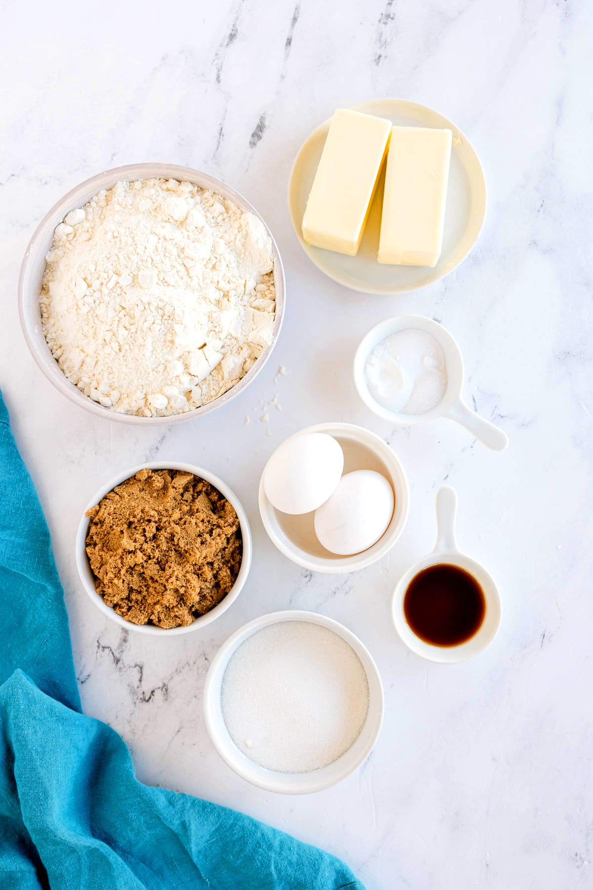 Bowls and plates with flour, unsalted butter, baking powder, baking soda, salt, sugar, vanilla extract, and eggs arranged on a white marble surface with a blue cloth. These are ingredients for Brown Butter Cookies