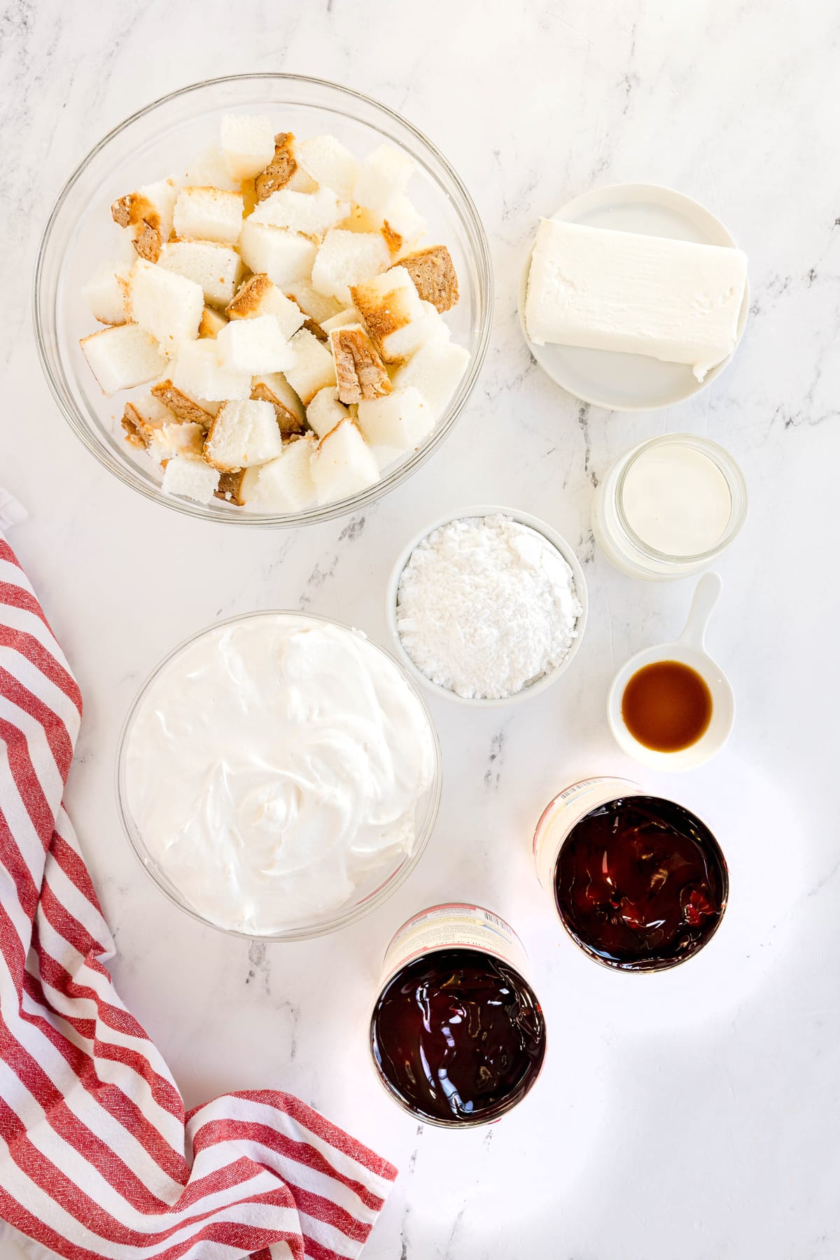 Top-down view of Ingredients for Cherries in the Snow are the following: Angel food cake, cream cheese, confectionaire's sugar, whole milk, vanilla extract, whipped topping and cherry pie filling with stripped linen on the side.