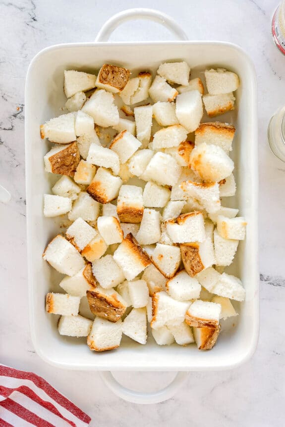 Cubed pieces of angel food cake are spread evenly in a white baking dish on a marble surface. This is first step in preparing Cherries in the Snow