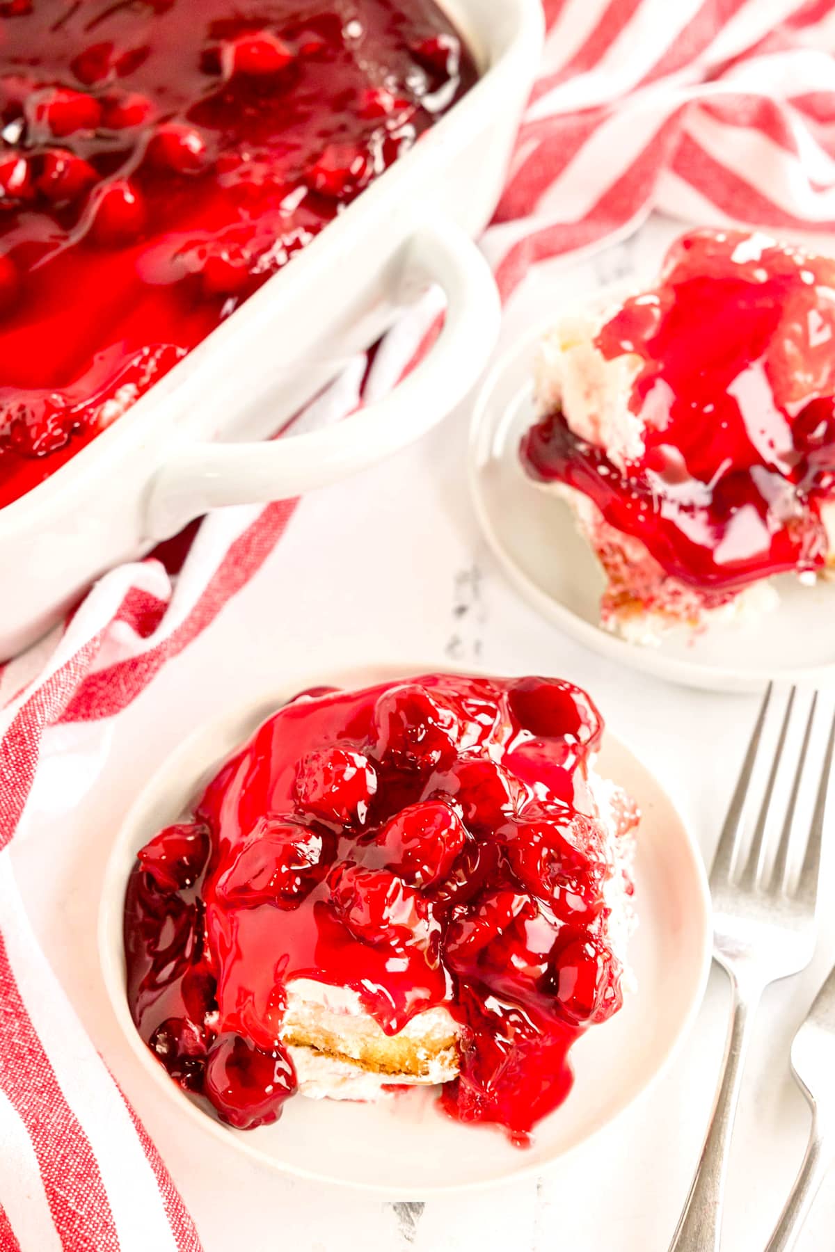 A serving of Cherries in the Snow with whipped topping on a plate, next to a larger dish of the same dessert, with a fork and a red-striped napkin nearby.