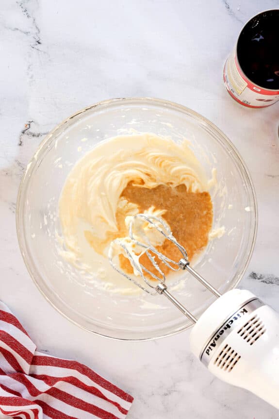 Next step in making Cherries in the Snow is to use a handheld electric mixer blending beat the softened cream cheese and confectioner’s sugar in a glass bowl on a marble countertop, with a striped towel and a bottle nearby.