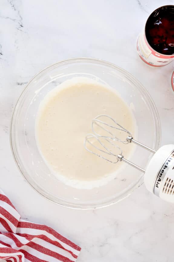 A glass bowl with milk and vanilla extract mixture is being blended with a handheld electric mixer on a marble surface. A red-striped towel and opened cans are nearby. This is one of the steps in preparing Cherries in the Snow