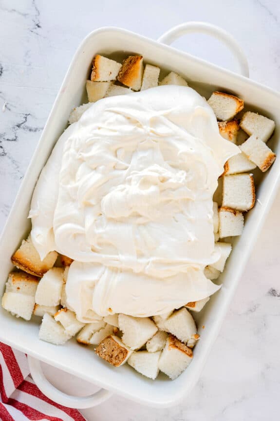 Cubed pieces of angel food cake are arranged in a white baking dish and topped with a thick layer of whipped cream or frosting for Cherries in the Snow recipe