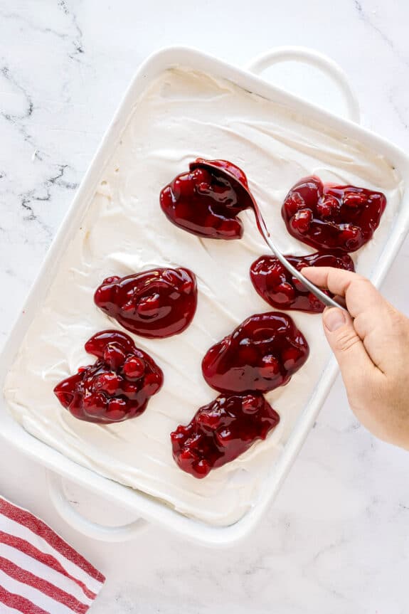 A hand spreads cherry pie filling over a layer of cream in a white rectangular baking dish on a marble surface. This is one of the steps in preparing Cherries in the Snow
