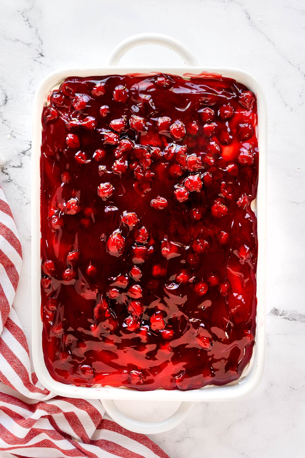 A rectangular white baking dish filled with dessert topped with glossy red cherry pie filling, placed on a white marble surface next to a red and white striped cloth for Cherries in the Snow recipe.