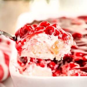 A spoon lifts a serving of Cherries in the Snow with layers of whipped cream, sponge cake, and cherry topping from a white baking dish.