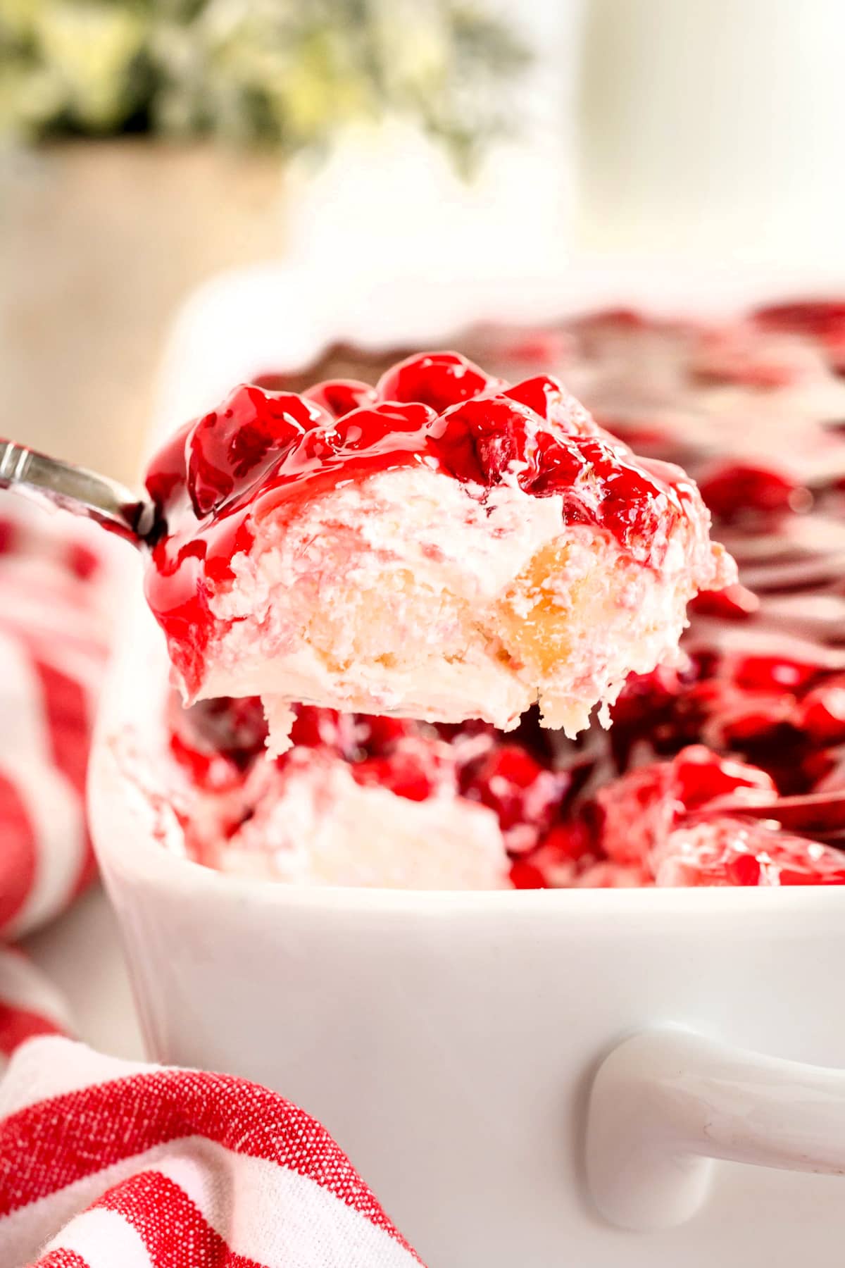 A close-up of a serving of Cherries in the Snow with whipped topping and cake layers being lifted from a white baking dish.