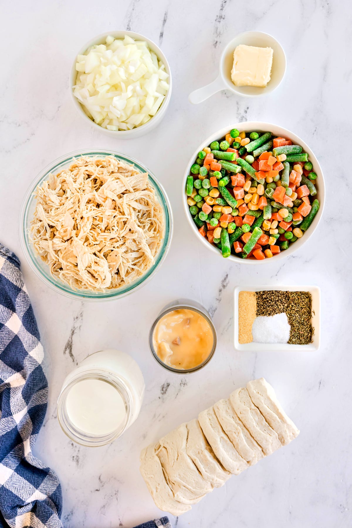 Overhead view of ingredients for Chicken Pot Pie Casserole: butter, onion, frozen mixed vegetables, cooked chicken, cream of chicken soup, milk, dried thyme, salt, black pepper, garlic powder, refrigerated biscuit dough. on a marble surface.