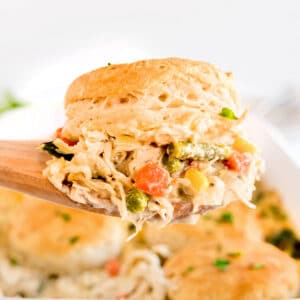 A wooden spoon holds a serving of creamy chicken and vegetable mixture topped with a golden biscuit, with more biscuits and filling visible in the background.