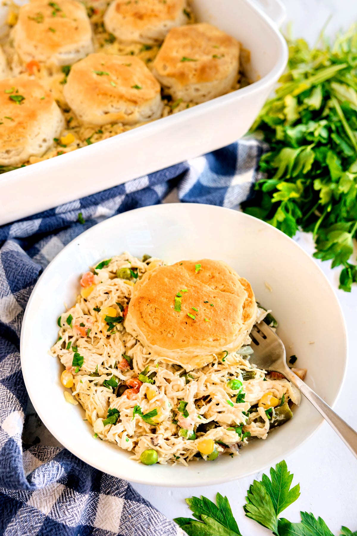 A white bowl filled with Chicken Pot Pie Casserole, vegetables, and topped with a biscuit, placed next to a baking dish of biscuits on a blue and white towel.