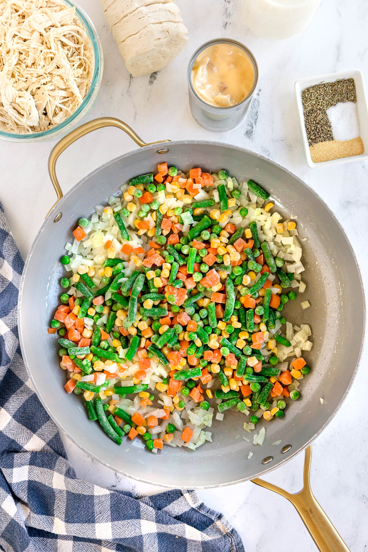 A skillet with sautéed chopped onions and frozen mixed vegetables on a marble countertop, surrounded by shredded chicken, a can of soup, biscuits, and seasonings for Chicken Pot Pie Casserole recipe