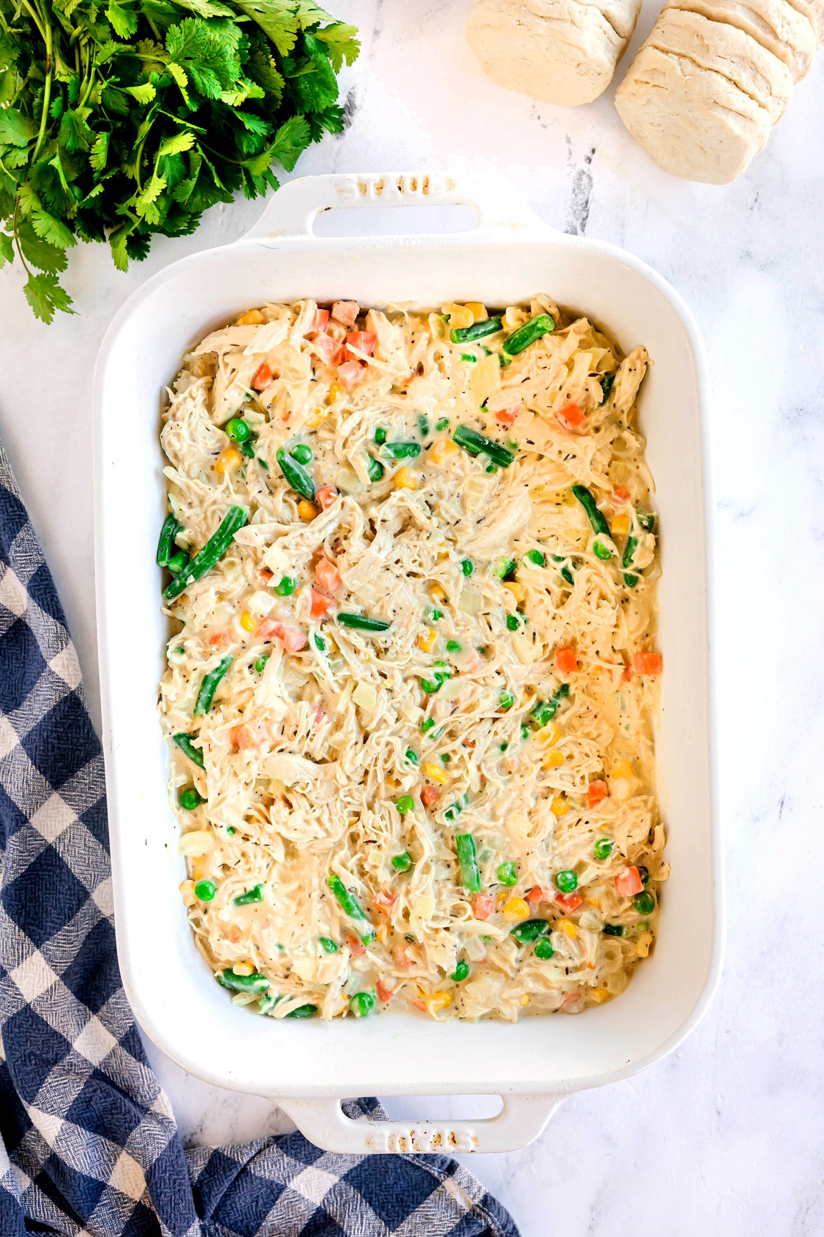 A white baking dish filled with creamy Chicken Pot Pie Casserole mixed with peas, carrots, and green beans, next to fresh parsley, biscuit dough, and a blue checkered cloth.