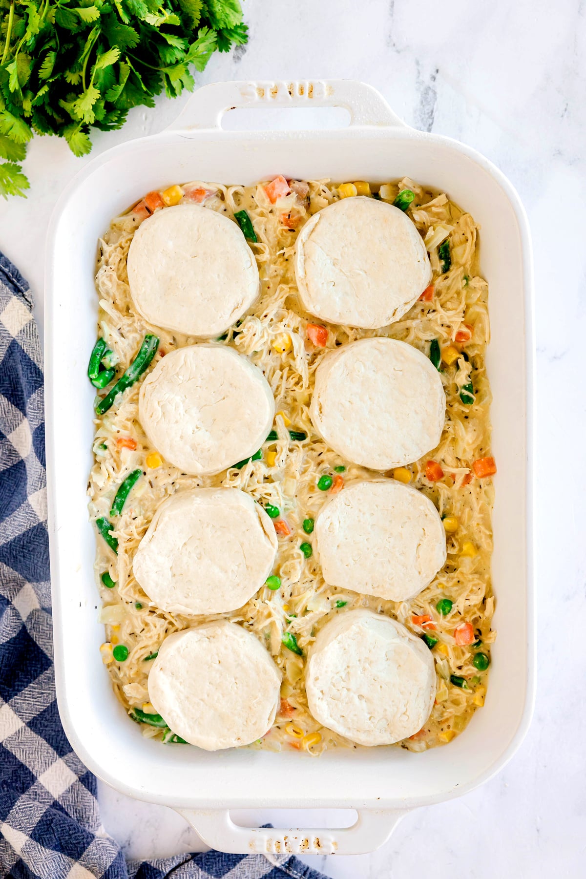 A white baking dish filled with creamy mixed vegetables and shredded chicken, topped with eight uncooked biscuit dough rounds, sits on a marble surface next to a checkered towel and fresh herbs. This is for Chicken Pot Pie Casserole recipe.