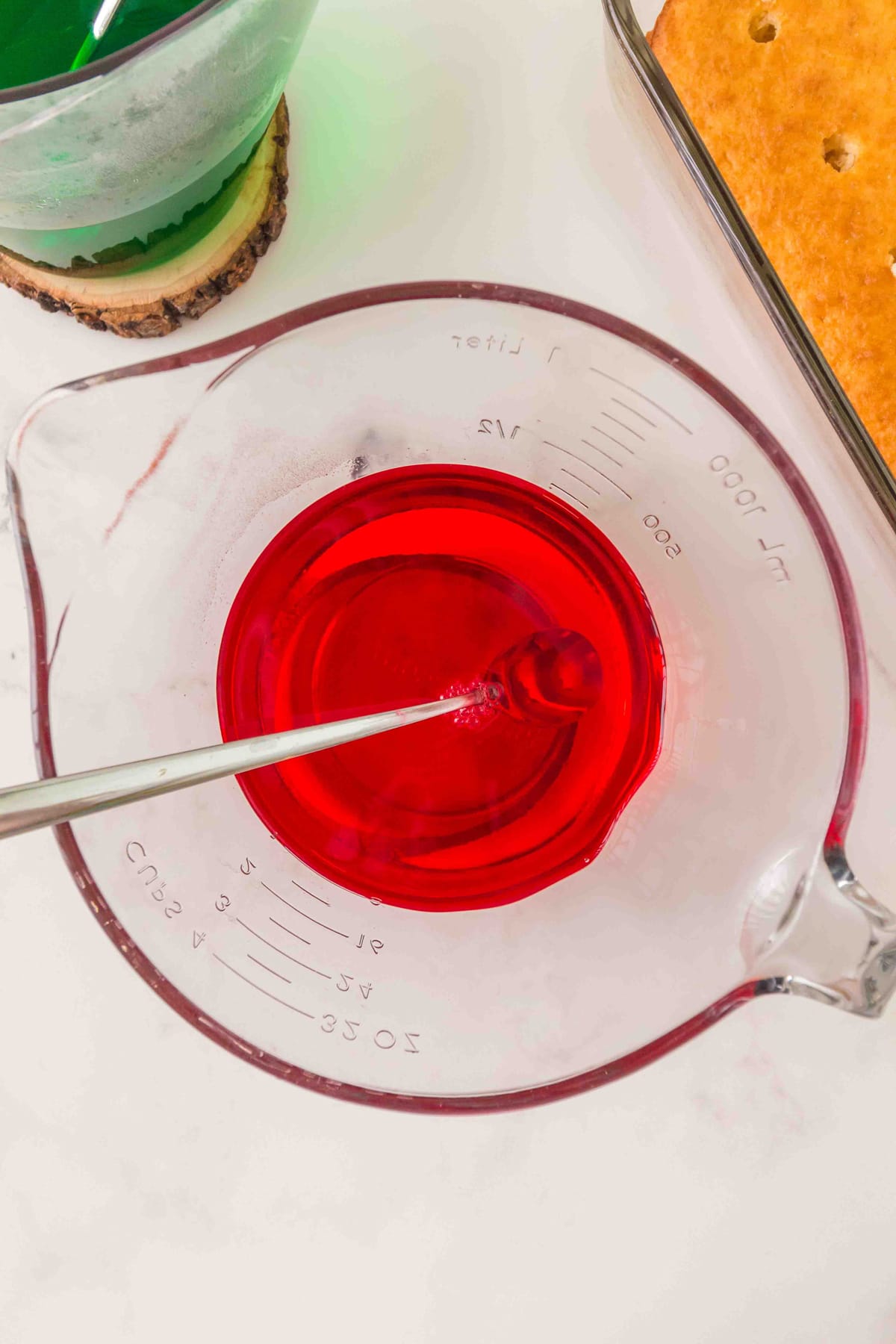 Preparing the jello for Christmas Poke Cake. A glass measuring cup filled with red Jell-o being stirred with a spoon, next to a green liquid jell-o in a cup and a baked cake in a glass dish.