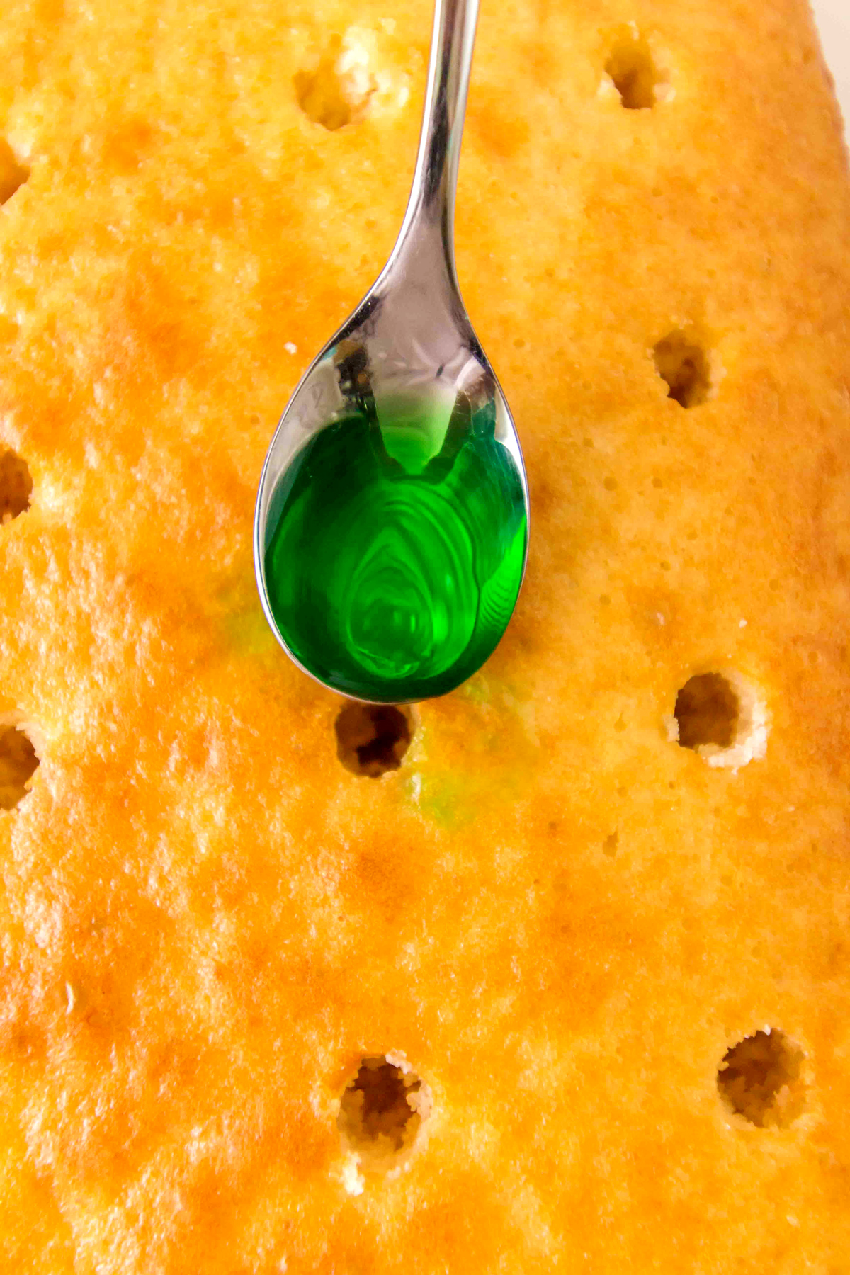Next step in preparing Christmas Poke Cake is to spoon the Jell-O mixtures into the holes, alternating colors for a festive pattern.